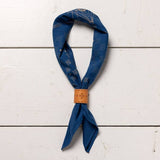 The Jenni Earle "Hell Yeah" bandana features a blue western pattern and a light brown wooden slider, displayed on a white wooden surface.
