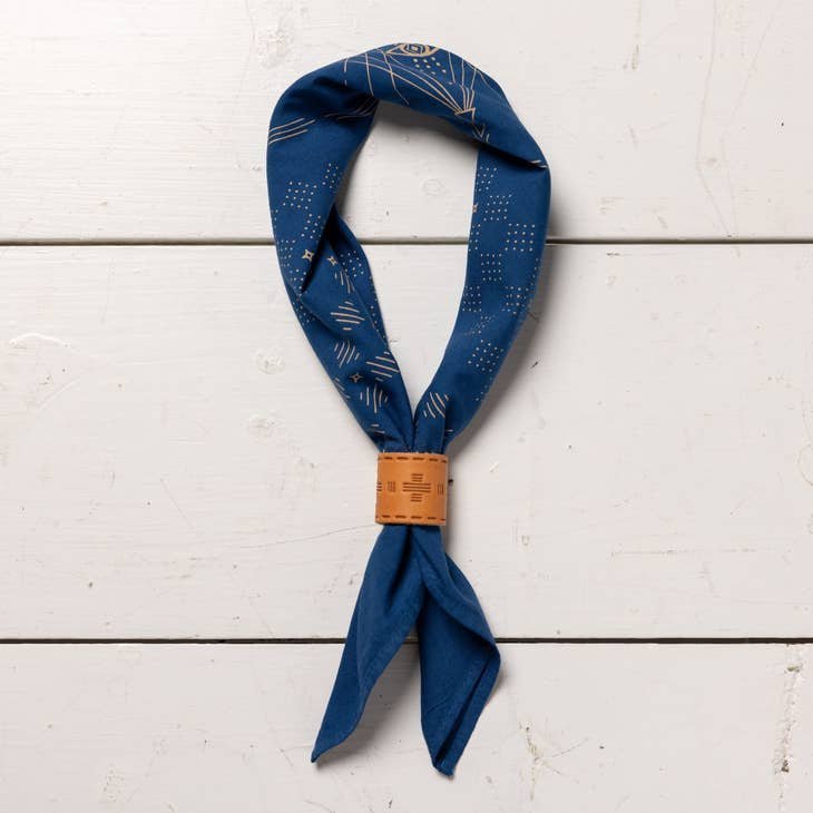 The Jenni Earle "Hell Yeah" bandana features a blue western pattern and a light brown wooden slider, displayed on a white wooden surface.