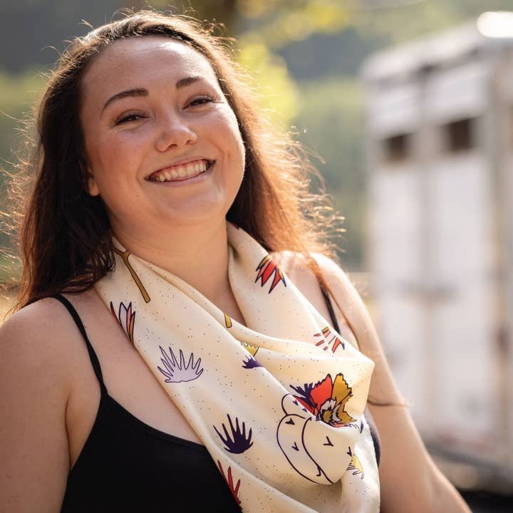 A woman with long brown hair smiles outdoors, wearing a black tank top and the Jenni Earle “I am Free Cactus” bandana—a vibrant, hand-illustrated piece that makes a perfect Valentine’s gift.