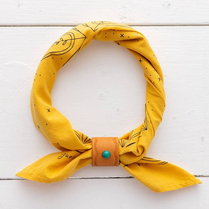 The Jenni Earle "Just Breathe" bandana features hand-dyed yellow fabric with black designs, rolled and secured by a tan leather snap closure with a turquoise button, displayed on a white wooden surface.
