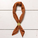 The Jenni Earle "Keep Going" Bandana features a rust hue with white dots and black geometric patterns, hand dyed for uniqueness—a thoughtful, motivational gift displayed with a gold leaf clip on white wood.