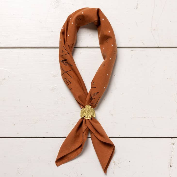 The Jenni Earle "Keep Going" Bandana features a rust hue with white dots and black geometric patterns, hand dyed for uniqueness—a thoughtful, motivational gift displayed with a gold leaf clip on white wood.