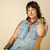 A person with a mullet, septum piercing, tattoos, and bracelets sits on a chair wearing a sleeveless denim shirt and the "Keep Going" bandana by Jenni Earle—a bold look that makes a great motivational gift.
