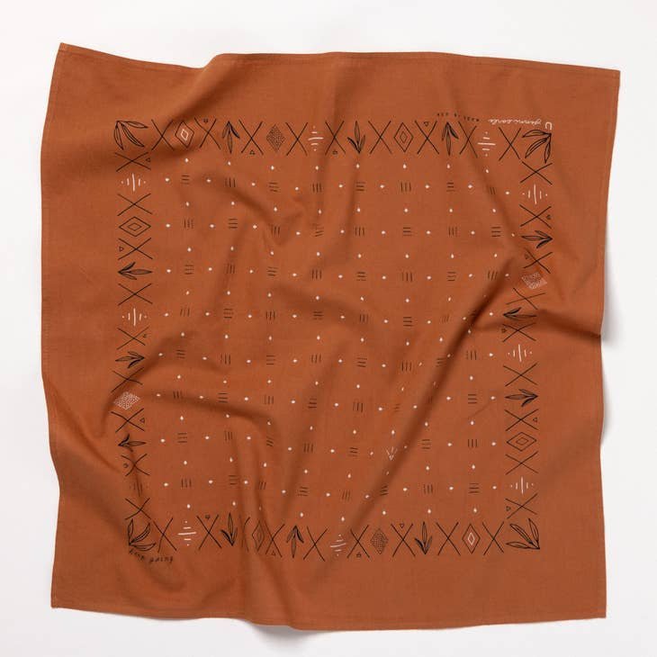The Jenni Earle "Keep Going" bandana is a rust-colored, hand-dyed accessory with black geometric patterns and border symbols—made in the USA and perfect as a motivational gift. Shown on a white background.