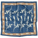 The "Kickin' Up Dust" bandana by Jenni Earle is a blue square scarf with a tan border and a white leafy botanical pattern, inspired by desert flowers.