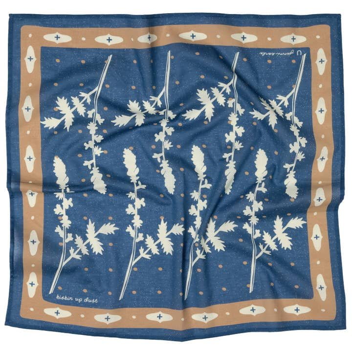 The "Kickin' Up Dust" bandana by Jenni Earle is a blue square scarf with a tan border and a white leafy botanical pattern, inspired by desert flowers.