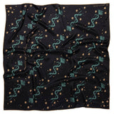 The "Lasso The Stars" bandana by Jenni Earle is a black square scarf with teal serpents, golden stars, and small dots—a magical silk accessory perfect for a distinctive holiday gift.