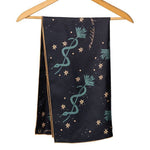 The "Lasso The Stars" bandana by Jenni Earle features green and gold star and plant motifs on black silk—an elegant holiday gift, shown hanging on a wooden hanger against a white background.