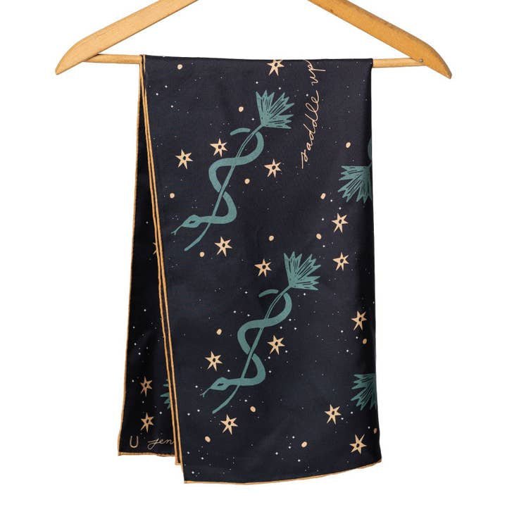 The "Lasso The Stars" bandana by Jenni Earle features green and gold star and plant motifs on black silk—an elegant holiday gift, shown hanging on a wooden hanger against a white background.