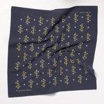 The Jenni Earle "Let Yourself Bloom" Bandana is a navy blue cotton square, featuring yellow and white floral motifs with small white dots. Slightly wrinkled, it's shown on a white background.