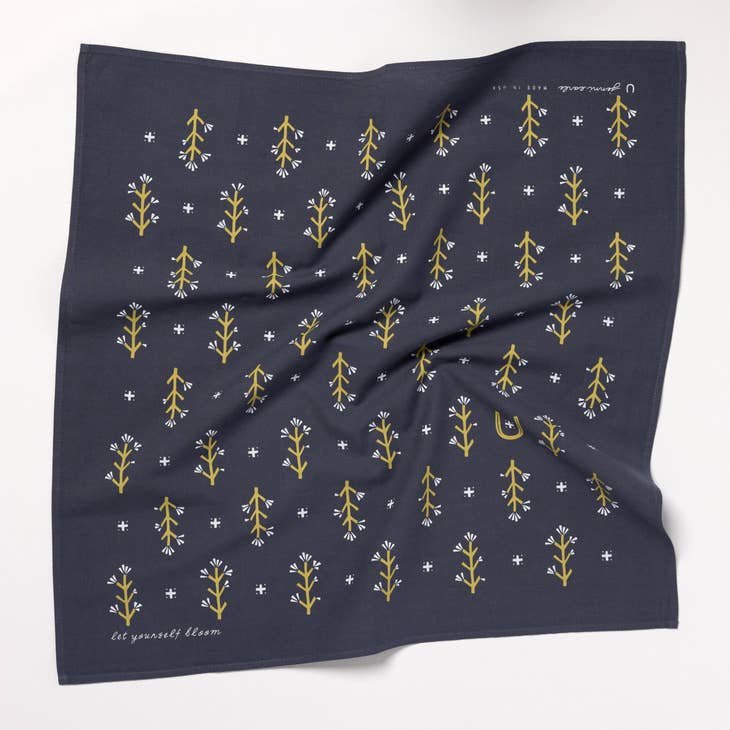 The "Let Yourself Bloom" Bandana by Jenni Earle is navy blue sustainable cotton, featuring yellow floral patterns, small white crosses, and the phrase "let yourself bloom" in one corner.
