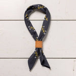 The “Let Yourself Bloom” bandana by Jenni Earle features a dark blue hand dyed fabric with yellow and white branch designs, tied with a tan leather ring and displayed on a white wooden surface.