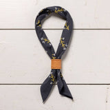 The Jenni Earle "Let Yourself Bloom" bandana features hand-dyed dark blue cotton with white and yellow branch designs. Folded and secured by a tan leather band, it’s displayed on a white wooden surface. Made from sustainable materials.