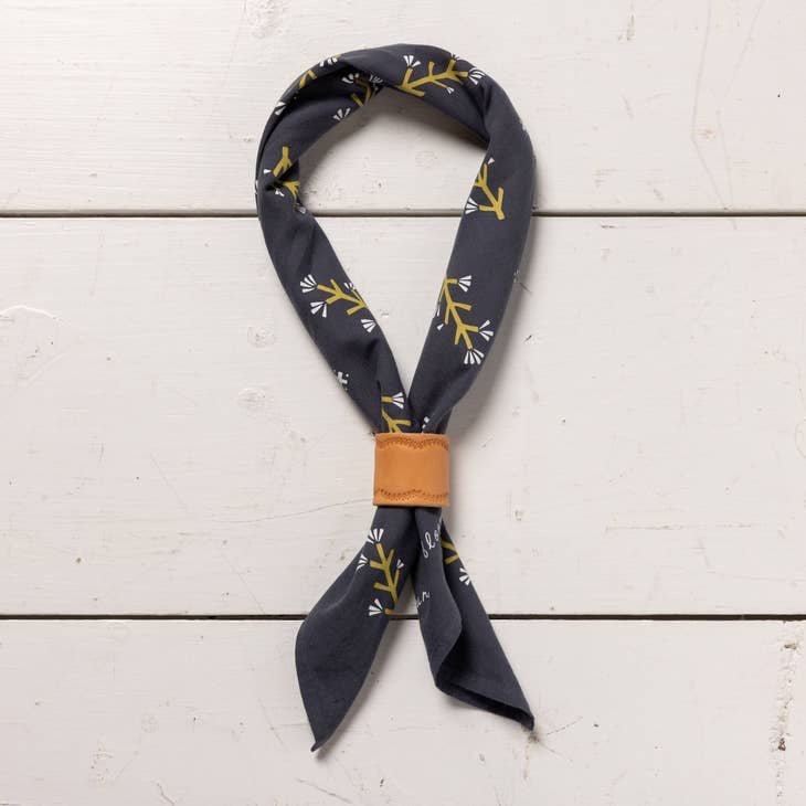 The “Let Yourself Bloom” bandana by Jenni Earle features a dark blue hand dyed fabric with yellow and white branch designs, tied with a tan leather ring and displayed on a white wooden surface.