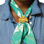 Wearing a denim shirt, someone styles the "Love Your Neighbor" Jenni Earle bandana as a green western scarf, secured with a gold thunderbird brooch.