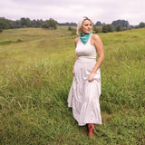 A woman in a sleeveless white top, long white skirt, red boots, and a Jenni Earle "Love Your Neighbor" Bandana stands in a grassy field with trees in the background.