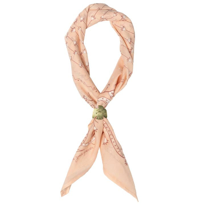 The Jenni Earle “Own Your Power” bandana is a light peach luxury cotton accessory featuring a subtle line pattern, tied in a loose knot and finished with a green decorative accent.