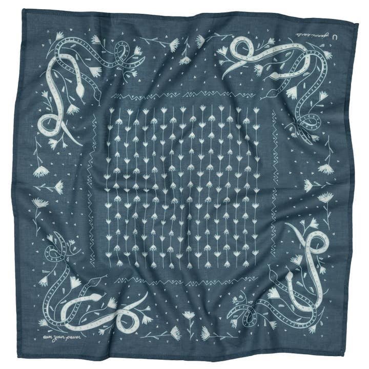 The "Own Your Power" Bandana by Jenni Earle is square and blue, featuring white dandelions at the center and stylized snakes and gladiolus in each corner, finished with a patterned border—ideal for expressing personal power.