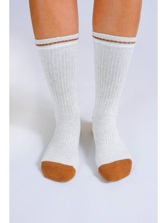A person stands on a white surface wearing Basal Crew Socks by Tailored Union: white cotton blend crew socks with brown toes, a reinforced heel and toe, and a brown stripe near the top.
