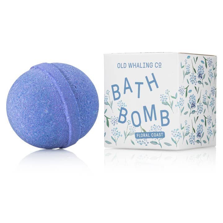 A purple floral bath bomb with hydrangea scent rests beside a box labeled “Bath Bomb | Floral Coast | Old Whaling Co.” The packaging features blue floral designs and the Old Whaling Co. brand name.