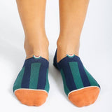 A pair of feet in Tailored Union Beholder Melon No Show Socks, made from premium combed cotton with navy and green stripes and orange toe caps, stands on a white surface.
