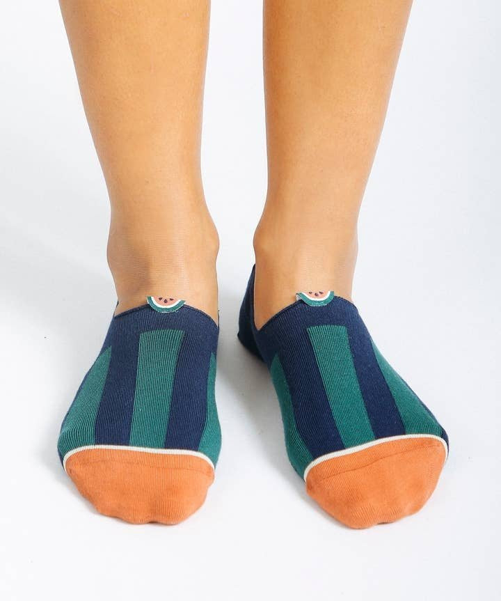 A pair of feet in Tailored Union Beholder Melon No Show Socks, made from premium combed cotton with navy and green stripes and orange toe caps, stands on a white surface.