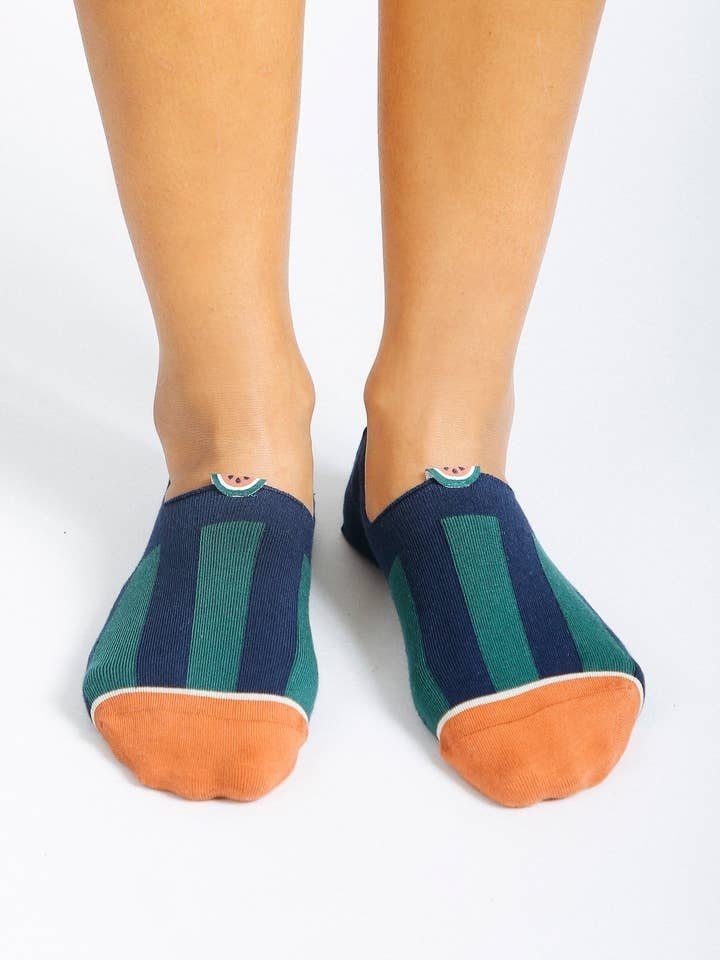 A pair of feet in Tailored Union Beholder Melon No Show Socks, made from premium combed cotton with navy and green stripes and orange toe caps, stands on a white surface.