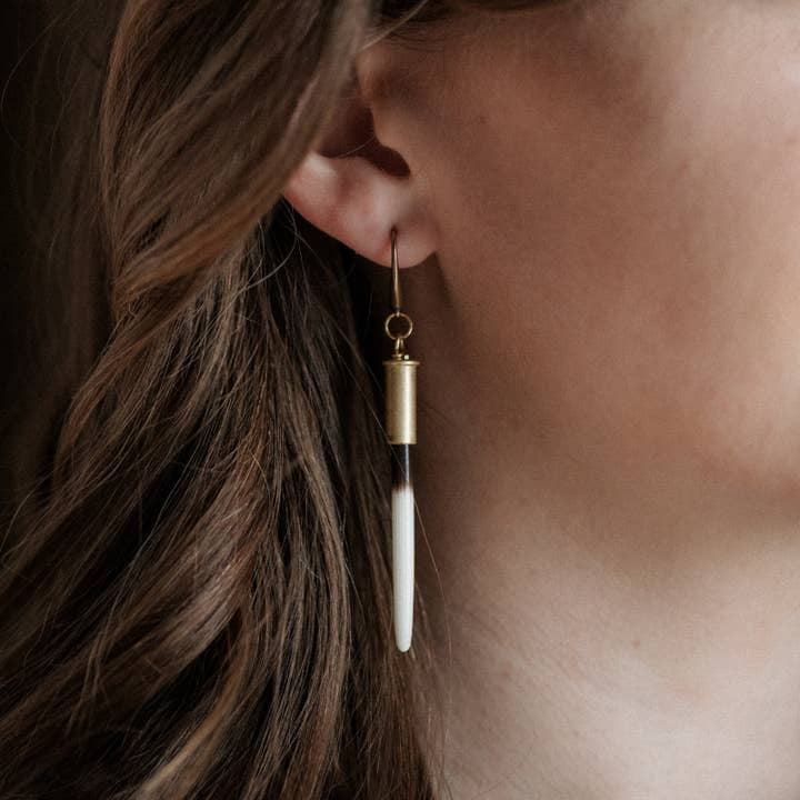 Close-up of a person wearing Beth Dutton Quill Earrings by CommonForm—long gold and white porcupine quills with brass ear hooks, partially hidden by wavy brown hair.