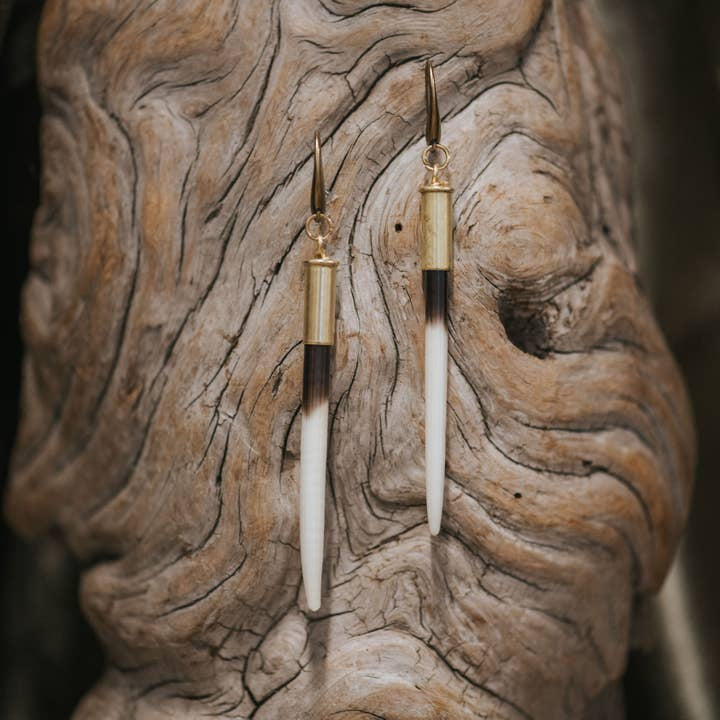 The Beth Dutton Quill Earrings by CommonForm feature long, pointed porcupine quills with gold and black accents, elegantly displayed against a textured wooden background.