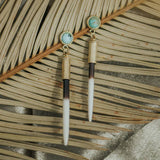 The Beth Dutton Turquoise Quill earrings by CommonForm feature turquoise stones and long, quill-inspired pendants with stud tops, shown resting on dried palm leaves.