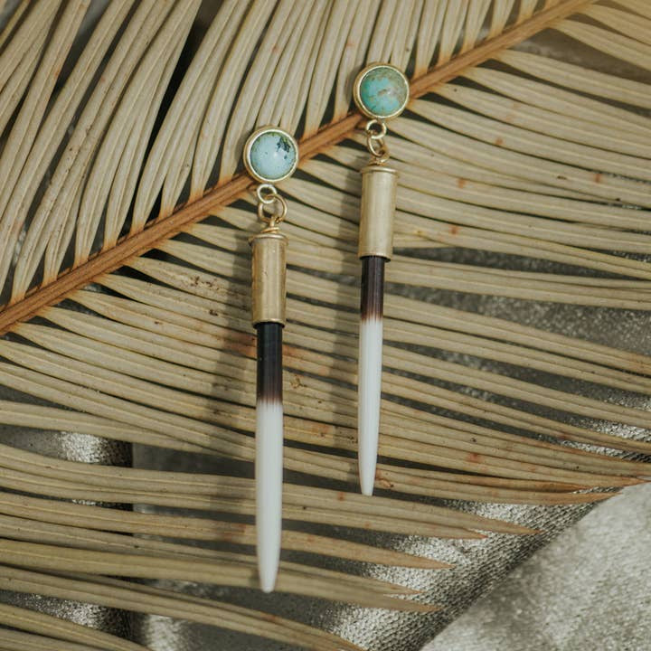 The Beth Dutton Turquoise Quill earrings by CommonForm feature turquoise stones and long, quill-inspired pendants with stud tops, shown resting on dried palm leaves.