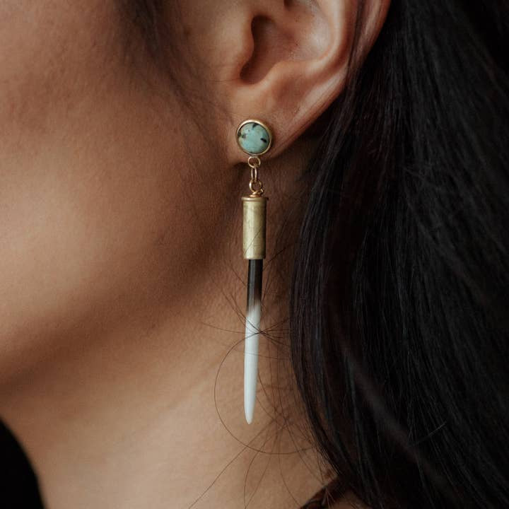 Close-up of a person wearing the Beth Dutton Turquoise Quill earring by CommonForm—a striking gold piece featuring a round turquoise stud and long, pointed pendant reminiscent of porcupine quills.
