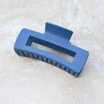The Tiepology Beyond the Border Hair Clip, in black and denim blue with a cutout center, rests on a light beige marble surface.