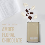 A bottle of Mozi’s Signature Cozy laundry detergent rests beside a white towel, surrounded by scattered chocolate pieces and flower petals. Text reads: “Smells like: Amber Floral Honey Chocolate.”.