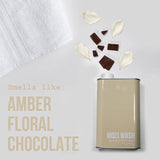 A bottle of Mozi’s Signature Cozy laundry detergent rests beside a white towel, surrounded by scattered chocolate pieces and flower petals. Text reads: “Smells like: Amber Floral Honey Chocolate.”.