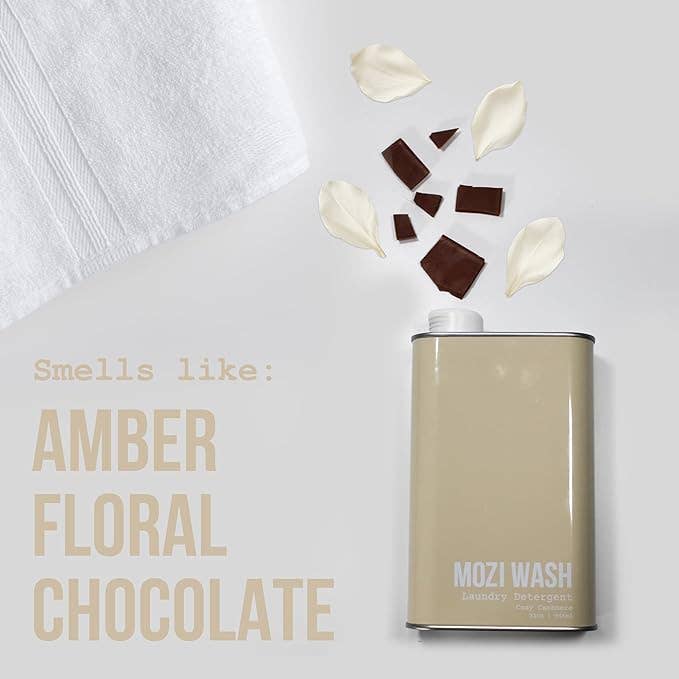 A bottle of Mozi’s Signature Cozy laundry detergent rests beside a white towel, surrounded by scattered chocolate pieces and flower petals. Text reads: “Smells like: Amber Floral Honey Chocolate.”.