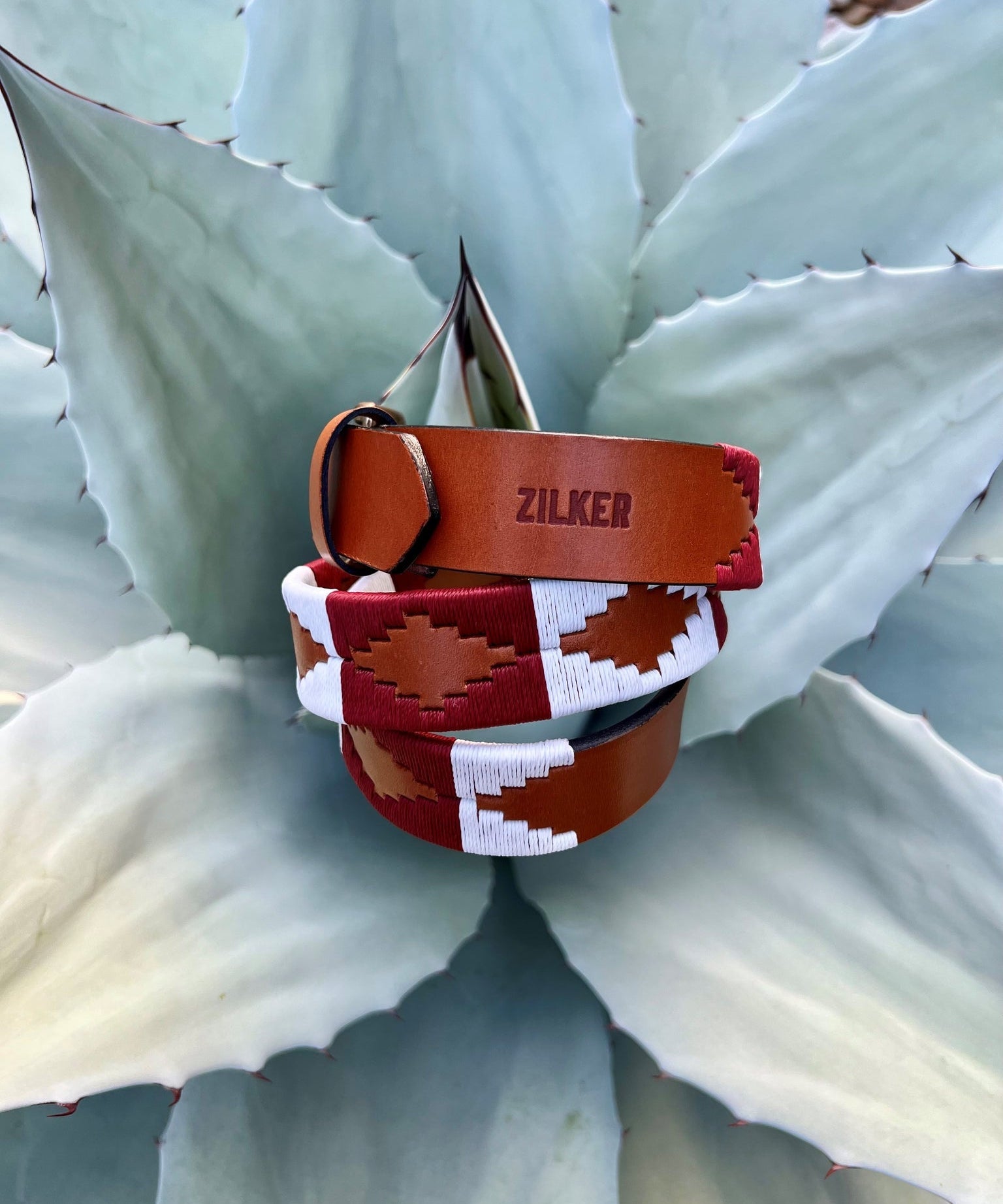 The Big Bend by Zilker Belts, crafted from hand-stitched Argentine leather and featuring a geometric "ZILKER" pattern, rests atop a pale green agave plant.