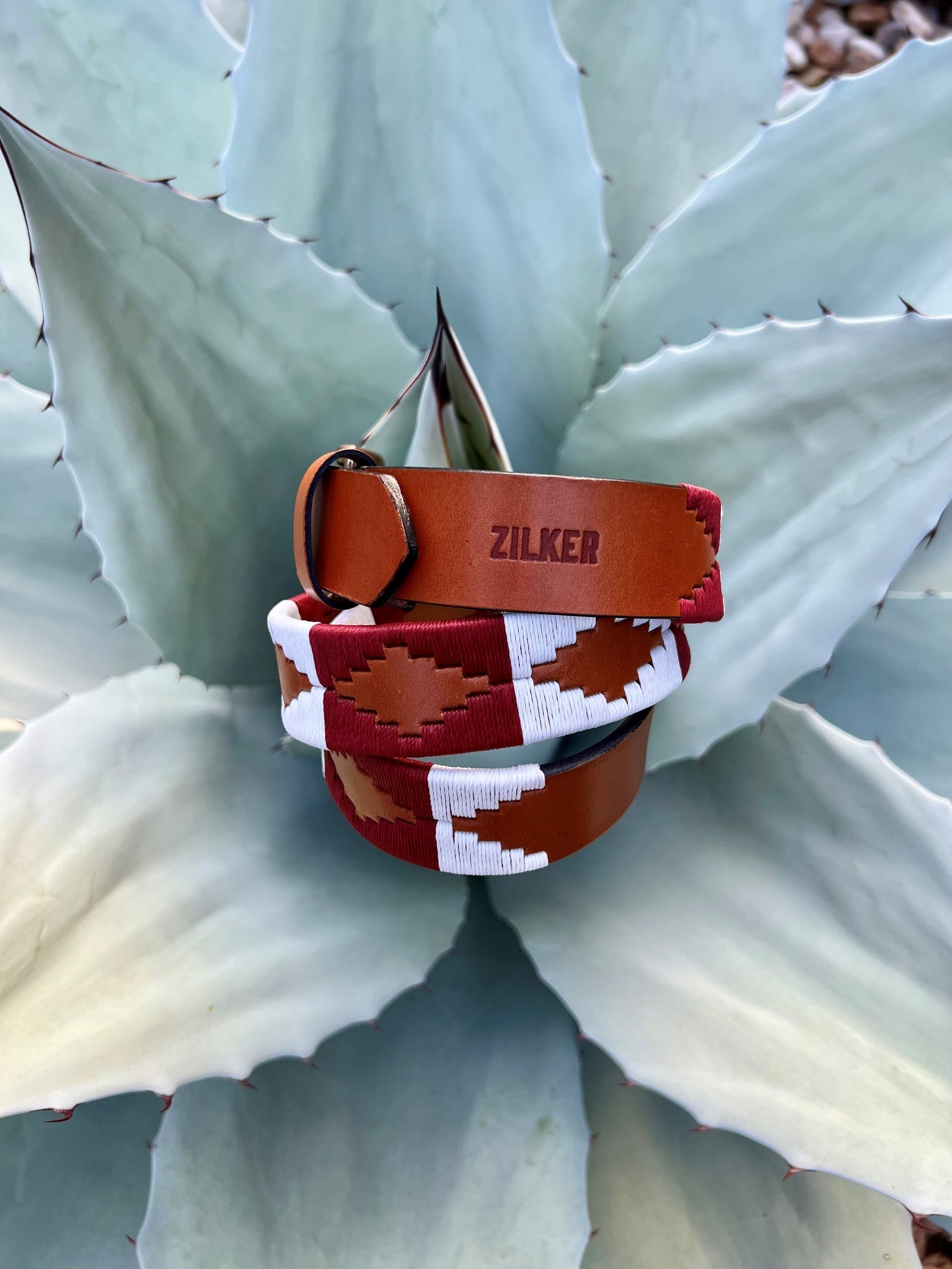 The Big Bend by Zilker Belts, crafted from hand-stitched Argentine leather and featuring a geometric "ZILKER" pattern, rests atop a pale green agave plant.