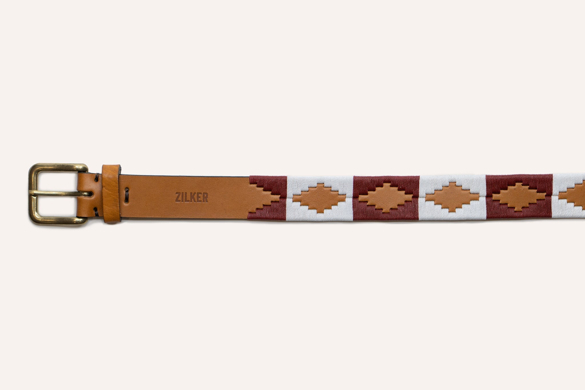 The Big Bend by Zilker Belts is a tan Argentine leather belt featuring a brass buckle, maroon and white woven geometric patterns, hand-stitched detailing, and "ZILKER" stamped on the belt.