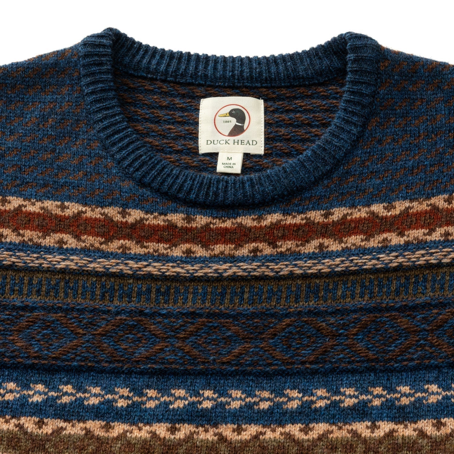 A close-up of the Duck Head Birch Fair Isle Crewneck Sweater features blue, brown, and tan geometric patterns and a Duck Head label at the neck. Machine washable.