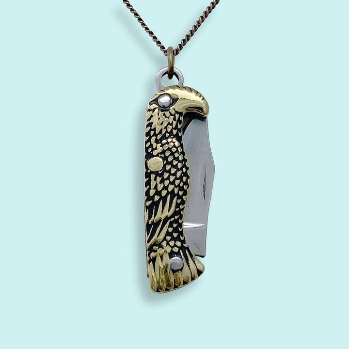 The Bird Knife Necklace by Ornamental Things features a pendant shaped like a bird’s head and body with a partially open stainless steel blade, hanging from a brass chain against a light blue background.