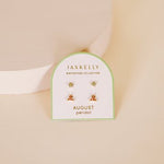 A display card labeled "Birthstone Earrings | JaxKelly" holds two pairs of August peridot birthstone earrings with 18K gold-plated sterling silver posts, shown on a light beige surface.