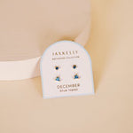A pair of JaxKelly Birthstone Earrings for December, featuring blue topaz and sterling silver posts, is displayed on a white card labeled "JaxKelly Birthstone Collection.