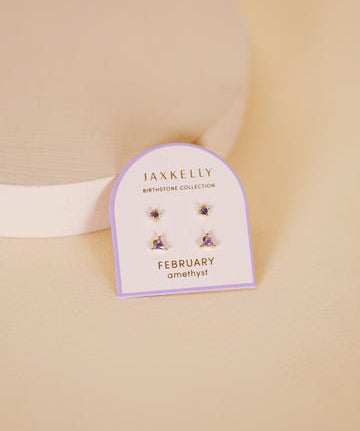 A pair of JaxKelly Birthstone Earrings featuring amethyst stones and hypoallergenic sterling silver posts rests on a branded card against a beige background.