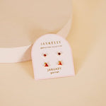 A card featuring JaxKelly Birthstone Earrings for January—small garnet studs with 18K gold plating and sterling silver posts—is displayed on a round platform against a soft beige background.