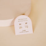 A pair of JaxKelly Birthstone Earrings for June, featuring mother of pearl and sterling silver posts, is displayed on a branded card against a beige background.
