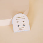The JaxKelly Birthstone Earrings for March feature aquamarine stones on sterling silver posts with 18K gold plating, presented on a branded card against a beige backdrop.