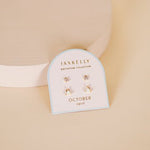 A pair of JaxKelly Birthstone Earrings for October, featuring opal stones on sterling silver posts, is shown on a small card against a light beige background.