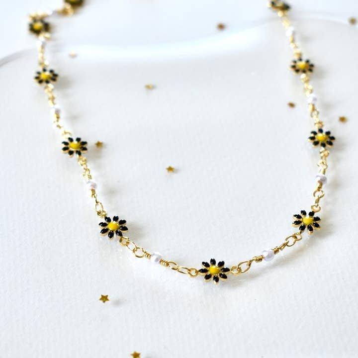 The Black Daisy Flower Pearl Chain Necklace by Vintage Acorn features gold plating, black and yellow daisy charms, and white beads, evoking 90's summer vibes. Shown on a white surface with gold star confetti.