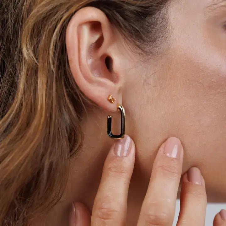 Close-up of a woman's ear wearing JaxKelly's Black Enamel Large Rectangle Gold Hoop earring. Her manicured hand touches her earlobe, highlighting the tarnish-free, geometric gold jewelry.